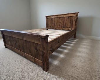 Op de afbeelding: Een rustiek houten bedframe met een hoofdbord en voetbord. Het bedframe is gemaakt van donkerbruin hout en heeft een lattenbodem. Het bedframe is leeg en staat op een beige tapijt. Afmetingen: ongeveer 200 cm lang en 150 cm breed.