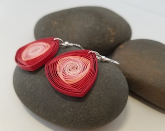 May include: A pair of red and pink paper quilled earrings with silver hooks. The earrings are shaped like a teardrop and have a spiral design in the center.