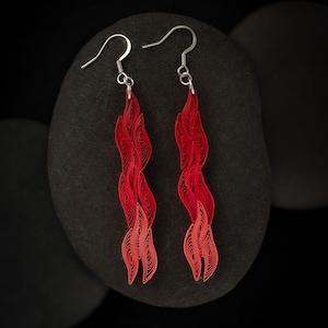 May include: A pair of red paper quilled earrings with silver hooks. The earrings are made of multiple layers of paper that have been rolled and shaped into a spiral design.