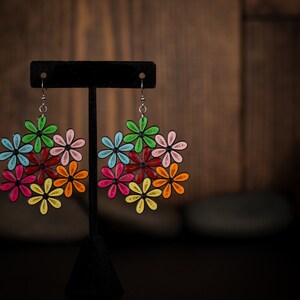 May include: Colorful floral earrings with a black outline. Each earring features a cluster of flowers in various colors, including green, blue, red, pink, orange, and yellow. The earrings are displayed on a black stand against a blurred wooden background.