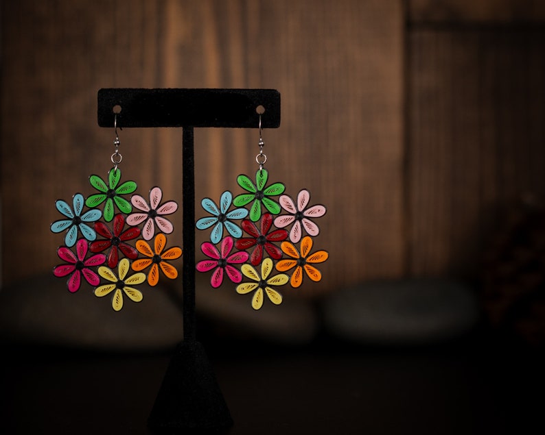 May include: Colorful floral earrings with a black outline. Each earring features a cluster of flowers in various colors, including green, blue, red, pink, orange, and yellow. The earrings are displayed on a black stand against a blurred wooden background.