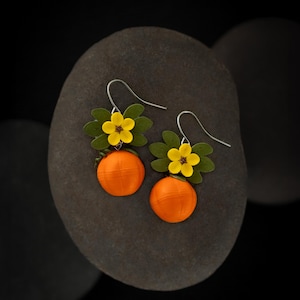 Puede incluir: Un par de pendientes colgantes de color naranja y amarillo con un diseño de flor y fruta. Los pendientes están hechos de arcilla polimérica y tienen ganchos de plata.