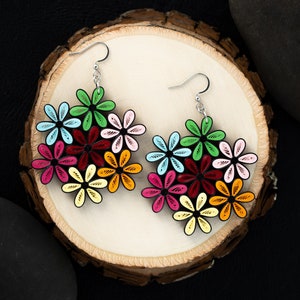 May include: A pair of colorful flower earrings. Each earring features a cluster of layered, daisy-like flowers in shades of red, pink, yellow, green, and blue. The earrings are set against a light wood slice background.