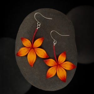 Puede incluir: Un par de pendientes de color naranja y rojo en forma de flor con ganchos plateados. Los pendientes están hechos de cuero y tienen un diseño delicado e intrincado.