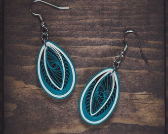 May include: A pair of teal and white paper earrings with a delicate, intricate design. The earrings are oval-shaped and have a silver hook.