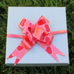 May include: A small, light gray gift box with a bright pink ribbon and bow. The bow features a heart pattern. The box is sitting on a bed of green grass.