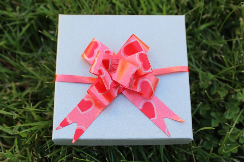 May include: A small, light gray gift box with a bright pink ribbon and bow. The bow features a heart pattern. The box is sitting on a bed of green grass.