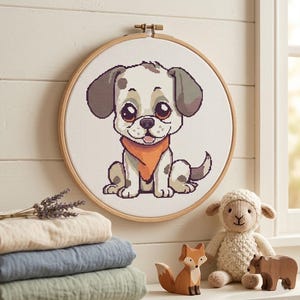 Peut inclure: Une broderie au point de croix d'un chiot de dessin animé avec des oreilles, des taches et un bandana marron, placée dans un cercle en bois. Le chiot est entouré d'une pile de tissu plié, d'un renard en bois, d'un agneau en peluche et d'un ours en bois.