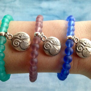 Puede incluir: Tres pulseras con dijes de plata en forma de concha. Las pulseras están hechas de cuentas de vidrio esmerilado en tonos turquesa, lavanda y azul.