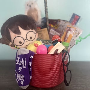 May include: A red plastic basket filled with various items including a plush character with glasses, a white owl figure, a purple sock with white text, a yellow rubber duck, a pink egg, a black wand, and black plastic round-rimmed glasses.