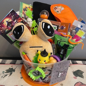 May include: A Halloween-themed gift basket filled with toys, candy, and accessories. The basket includes a plush toy with large black eyes, a Pikachu figurine wearing a witch hat, and various Halloween-themed treats and trinkets. The basket is decorated with a Halloween pattern.