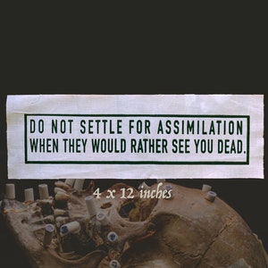 May include: A white paper sign with black text that reads "DO NOT SETTLE FOR ASSIMILATION WHEN THEY WOULD RATHER SEE YOU DEAD." The sign is torn and has a rough texture. The sign is placed on top of a human skull with a dark background. The skull is brown and has a few white teeth. The skull is 4 x 12 inches.