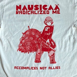 以下が含まれることがあります： 風立ちぬの谷のナウシカのアニメ映画のキャラクターの赤いグラフィックプリントが施された白いTシャツ。 「ナウシカは私を過激化した」と「共犯者は同盟国ではない」というテキストが赤色でTシャツに印刷されています。