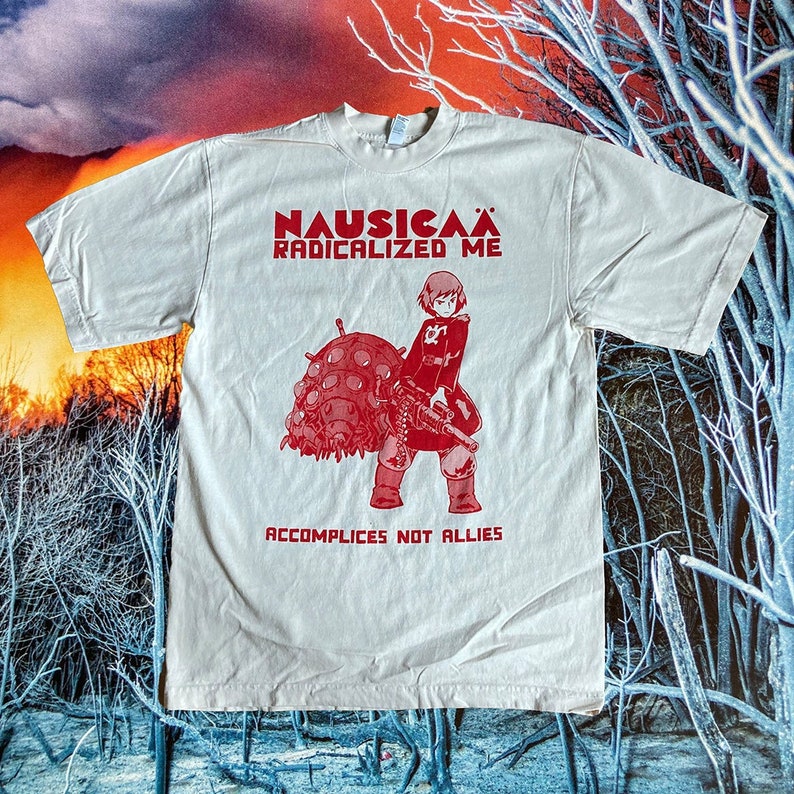 Puede incluir: Una camiseta blanca con un estampado gr&aacute;fico rojo de un personaje de la pel&iacute;cula de anime Nausica&auml; del Valle del Viento. El texto "Nausica&auml; Radicalized Me" y "Accomplices Not Allies" est&aacute; impreso en rojo en la camiseta.