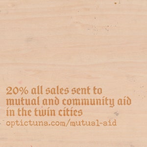Puede incluir: Un letrero de madera con el texto "20% de todas las ventas enviadas a la ayuda mutua y comunitaria en las ciudades gemelas optictuna.com/mutual-aid" en una fuente marr&oacute;n.