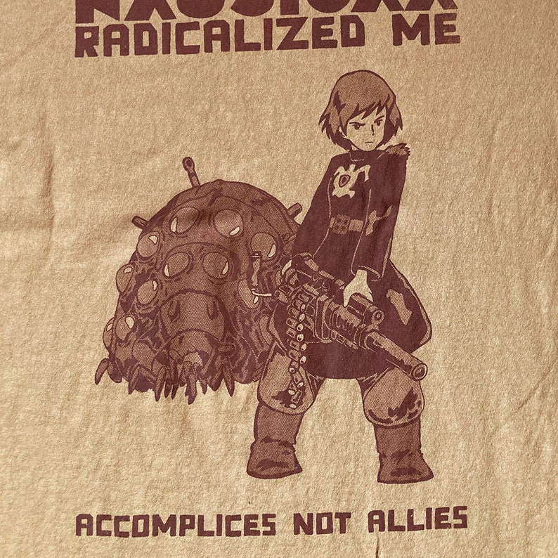 Puede incluir: Una camiseta marr&oacute;n con un gr&aacute;fico de una chica sosteniendo un arma y un insecto grande. El texto "RADICALIZED ME" est&aacute; por encima del gr&aacute;fico y "ACCOMPLICES NOT ALLIES" est&aacute; por debajo del gr&aacute;fico.
