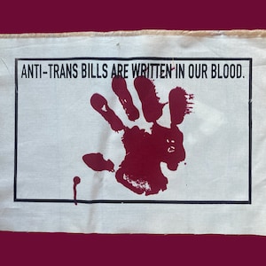 Może przedstawiać: Biały baner z tkaniny z czarnym tekstem, który brzmi "ANTI-TRANS BILLS ARE WRITTEN IN OUR BLOOD." Czerwony odcisk dłoni znajduje się w środku banera.