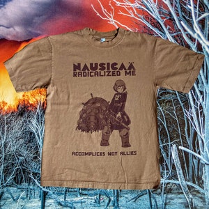 Puede incluir: Una camiseta marr&oacute;n con un estampado gr&aacute;fico de un personaje de la pel&iacute;cula de anime Nausica&auml; del Valle del Viento. El texto "Nausica&auml; Radicalized Me" y "Accomplices Not Allies" est&aacute; impreso en la camiseta.