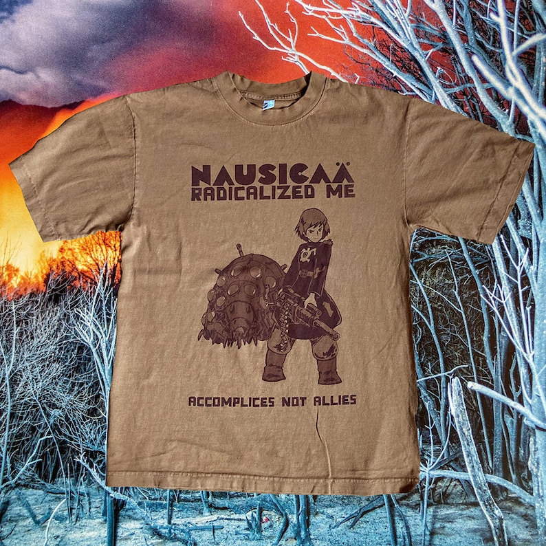 Puede incluir: Una camiseta marr&oacute;n con un estampado gr&aacute;fico de un personaje de la pel&iacute;cula de anime Nausica&auml; del Valle del Viento. El texto "Nausica&auml; Radicalized Me" y "Accomplices Not Allies" est&aacute; impreso en la camiseta.