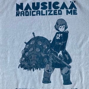 Puede incluir: Una camiseta gris claro con un estampado gr&aacute;fico azul de un personaje femenino sosteniendo una ametralladora y montando una criatura grande de m&uacute;ltiples ojos. El texto "NAUSICA&Auml; RADICALIZED ME" est&aacute; impreso sobre el gr&aacute;fico y "ACCOMPLICES NOT ALLIES" est&aacute; impreso debajo del gr&aacute;fico.
