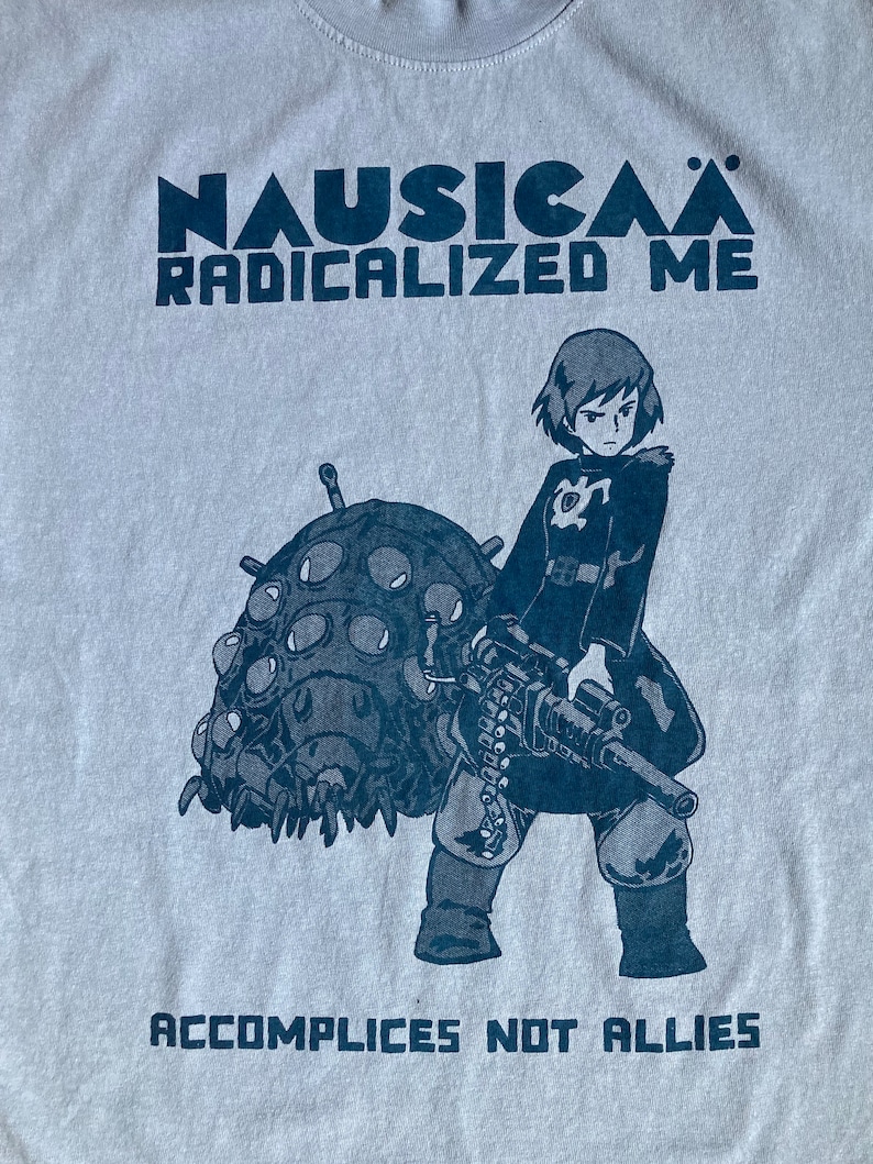 Puede incluir: Una camiseta gris claro con un estampado gr&aacute;fico azul de un personaje femenino sosteniendo una ametralladora y montando una criatura grande de m&uacute;ltiples ojos. El texto "NAUSICA&Auml; RADICALIZED ME" est&aacute; impreso sobre el gr&aacute;fico y "ACCOMPLICES NOT ALLIES" est&aacute; impreso debajo del gr&aacute;fico.