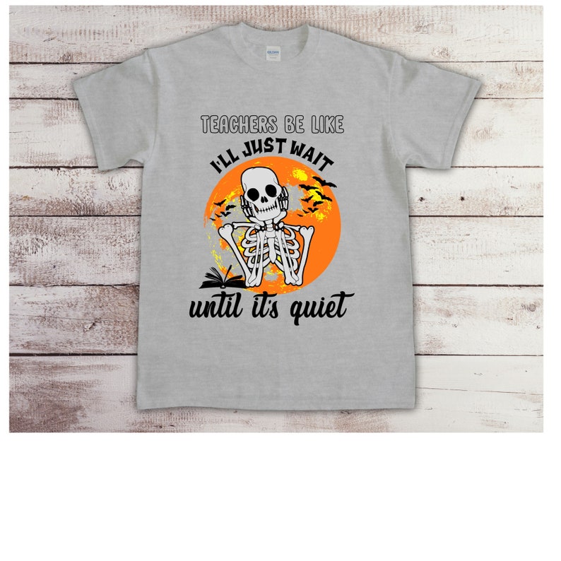 May include: A light gray t-shirt with the text "TEACHERS BE LIKE I'LL JUST WAIT until it's quiet." The graphic features a skeleton wearing headphones in front of an orange moon with bats. The skeleton is holding a book.