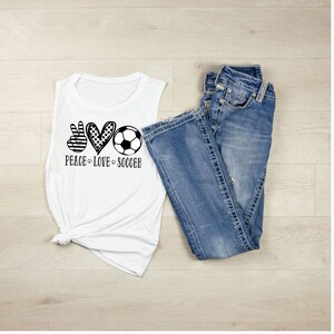 May include: White sleeveless muscle tank top with a black and white graphic of a peace sign, heart, and soccer ball. The text "Peace Love Soccer" is below the graphic.