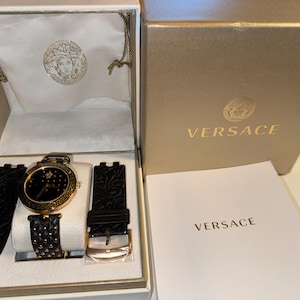 Puede incluir: Un reloj Versace negro y dorado con una correa de cuero negro y una hebilla dorada. El reloj est&aacute; en una caja blanca con un logotipo dorado de Versace, dentro de una caja dorada con el nombre Versace impreso.
