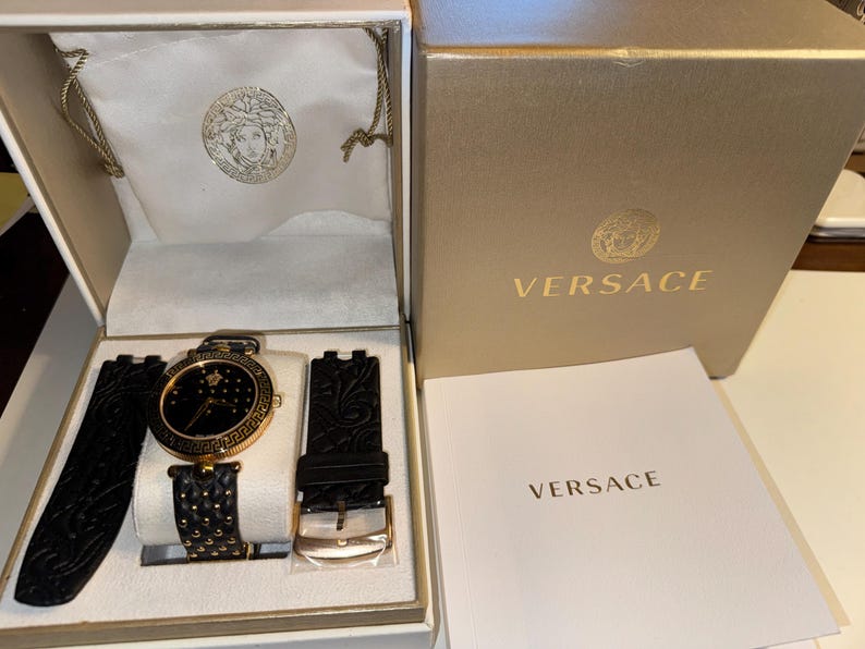 Puede incluir: Un reloj Versace negro y dorado con una correa de cuero negro y una hebilla dorada. El reloj est&aacute; en una caja blanca con un logotipo dorado de Versace, dentro de una caja dorada con el nombre Versace impreso.