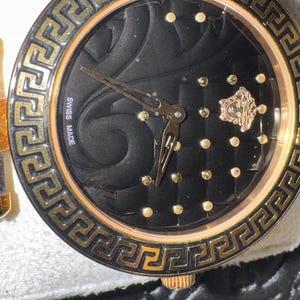 Puede incluir: Primer plano de un reloj de lujo con esfera negra y detalles dorados. El reloj presenta una esfera texturizada, marcadores de hora dorados y un bisel dorado con un dise&ntilde;o de greca. Las manecillas son doradas y el reloj est&aacute; marcado "SWISS MADE".