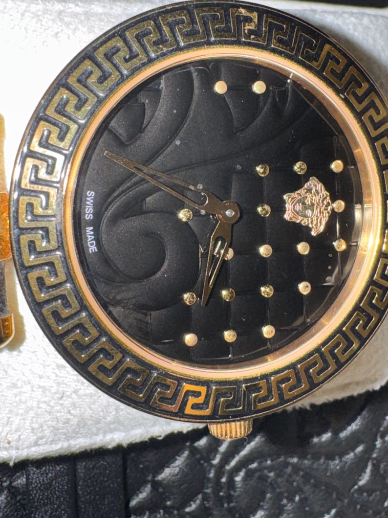 Puede incluir: Primer plano de un reloj de lujo con esfera negra y detalles dorados. El reloj presenta una esfera texturizada, marcadores de hora dorados y un bisel dorado con un dise&ntilde;o de greca. Las manecillas son doradas y el reloj est&aacute; marcado "SWISS MADE".