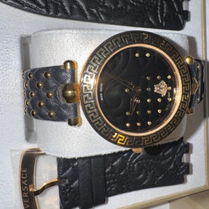 Puede incluir: Reloj de pulsera negro y dorado con una correa de cuero negro y una correa de repuesto. La esfera del reloj es negra con detalles dorados y un bisel dorado. El reloj tiene una hebilla dorada y la palabra "VERSACE" est&aacute; impresa en la correa.