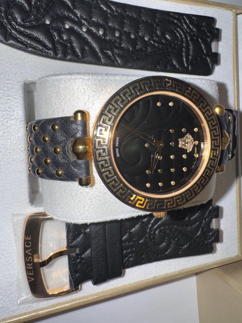 Puede incluir: Reloj de pulsera negro y dorado con una correa de cuero negro y una correa de repuesto. La esfera del reloj es negra con detalles dorados y un bisel dorado. El reloj tiene una hebilla dorada y la palabra "VERSACE" est&aacute; impresa en la correa.