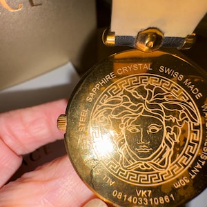 Puede incluir: Primer plano de un reloj dorado con un emblema de cabeza de Medusa. El reloj tiene una correa de cuero negro y un bisel dorado. El reloj muestra texto como "SAPPHIRE CRYSTAL", "SWISS MADE" y "WATER RESISTANT 30m".
