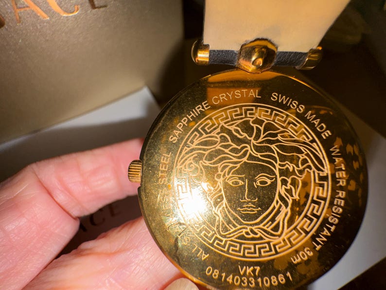 Puede incluir: Primer plano de un reloj dorado con un emblema de cabeza de Medusa. El reloj tiene una correa de cuero negro y un bisel dorado. El reloj muestra texto como "SAPPHIRE CRYSTAL", "SWISS MADE" y "WATER RESISTANT 30m".