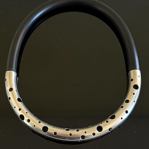 Könnte beinhalten: Eine moderne Halskette mit einem schwarzen, abgerundeten Band, das mit einem silberfarbenen, perforierten Metallteil verbunden ist. Der Metallteil hat zahlreiche kreisförmige Aussparungen, die ein einzigartiges Design schaffen. Die Halskette wird vor einem dunklen Hintergrund präsentiert.