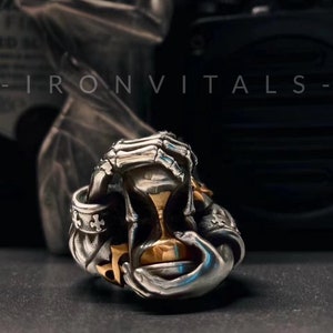 May include: A silver ring with a skeletal hand design holding an hourglass. The hourglass is made of gold and has a black background. The ring is set against a dark background.