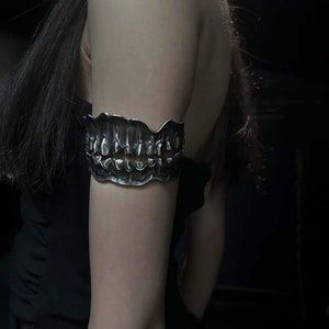 Puede incluir: Un brazalete plateado con forma de hilera de dientes. El brazalete tiene una superficie texturizada e irregular y se lleva en la parte superior del brazo. Los dientes están detallados, creando un accesorio audaz y original.