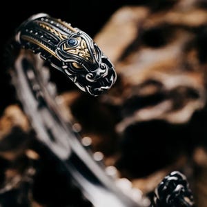 May include: A silver and gold bracelet with a detailed dragon head design. The dragon's eye is a blue gemstone.