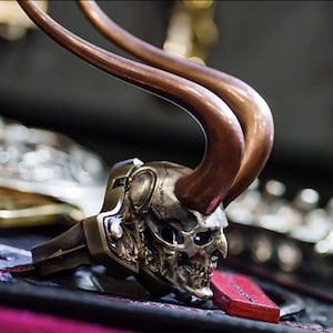 Puede incluir: Primer plano de un objeto con forma de calavera con extensiones curvas en forma de cuerno. La calavera es metálica con detalles intrincados, y los cuernos son de color bronce. El objeto parece ser una pieza decorativa o un accesorio.