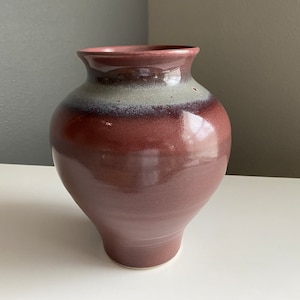 May include: A ceramic vase with a rounded body and a wide opening. The vase is glazed in a deep burgundy color with a band of light gray glaze around the middle.