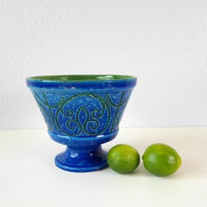 May include: A blue ceramic bowl with a green interior and decorative scrollwork. Two green limes sit beside the bowl. The bowl has a pedestal base and is likely vintage.