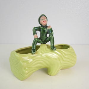 May include: A green ceramic planter shaped like a log with two separate planting areas. A green ceramic elf figurine sits on top of the log planter.