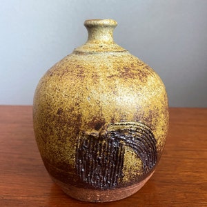 May include: A small, brown and yellow ceramic vase with a textured surface and a design of a stylized waterfall on the front.