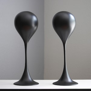 May include: Two matte black display stands, each featuring a smooth, spherical top section supported by a slender, elongated neck that flares out into a wide, circular base. These tall, sculptural objects have a uniform, solid dark finish.