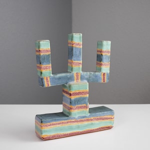 May include: A ceramic candle holder with a striped design in shades of blue, green, and pink. The holder has three arms and a rectangular base.