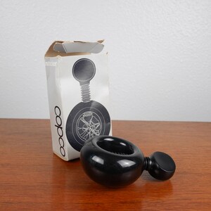 May include: Black plastic nutcracker shaped like a car wheel with a small knob on the side. The nutcracker is sitting on a wooden table next to a white box with the word "copco" printed on it.