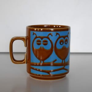 May include: Brown ceramic mug with a blue background featuring two brown owl illustrations. The mug has a brown handle and rim. A decorative item for the kitchen or home.