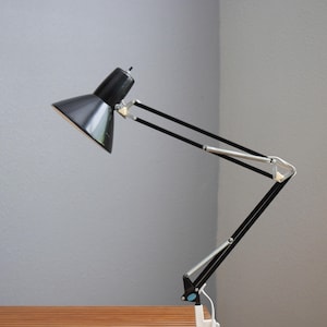 May include: A black articulated desk lamp with a conical shade and a multi-jointed arm. The lamp features silver-toned metal connectors and a white base clamp. The structure allows for adjustable positioning of the light head and arm segments.