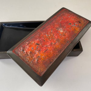 May include: A rectangular ceramic box with a lid. The lid is decorated with a red and orange glaze with a textured finish. The box is black.